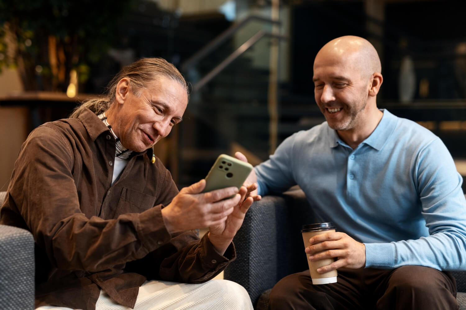 Homem sorridente mostra informações no celular para colega enquanto tomam café, ilustrando a verificação de fake news sobre FGTS na Facta Online.